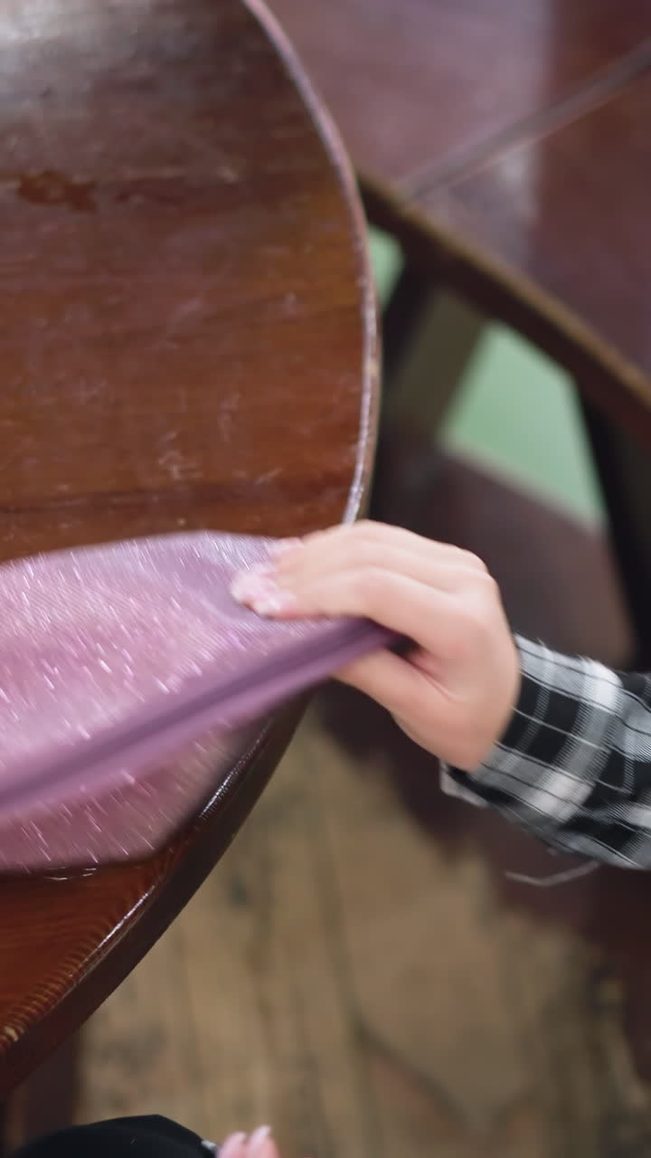 Hands pull sanitizer bottle from pouch beside mask and glasses on wooden table, plaid sleeve, careful unzipping and placement, clear liquid bottle, hygiene ritual for commuter, daylight cafe