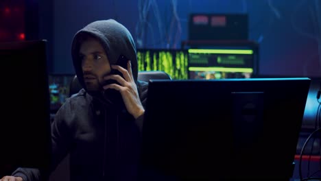 young man software developer and programmer in a hood working at night at the two big screen of computers in the dark room and speaking on the mobile phone