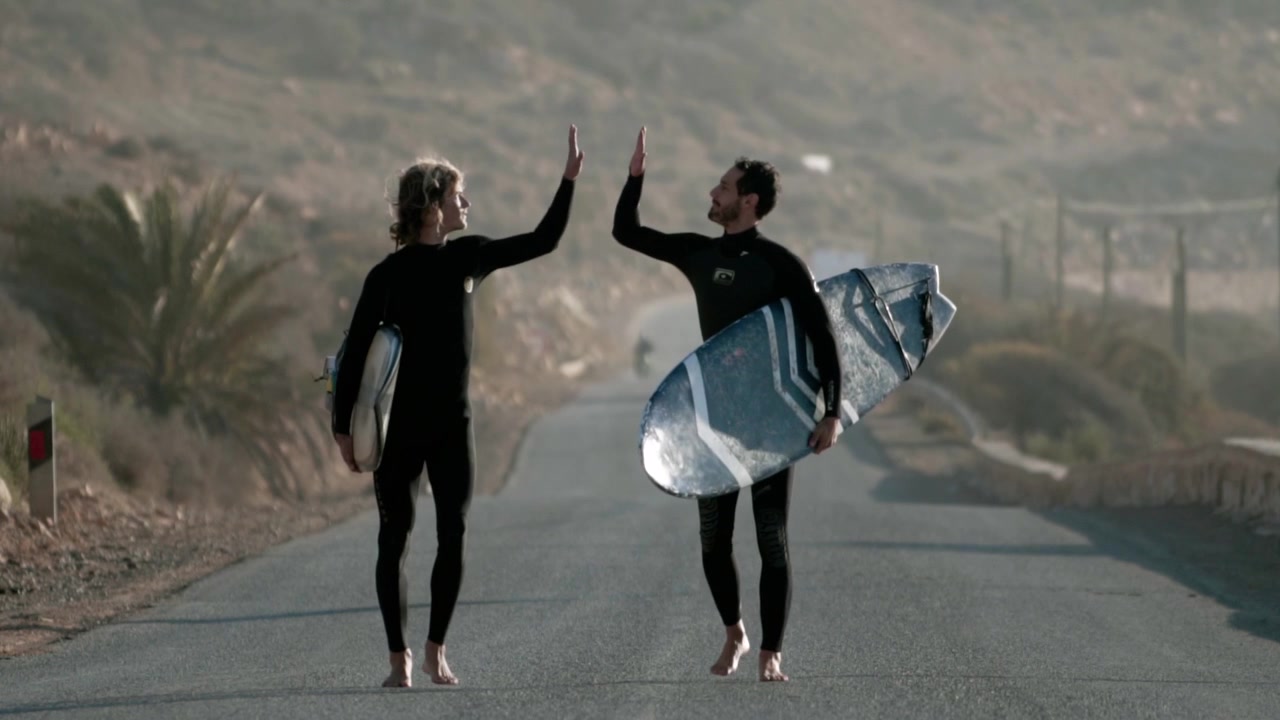 Dois surfistas caucasianos, vestindo roupa de neoprene e segurando suas pranchas de surf, caminham em uma estrada marroquina em direção à câmera durante o nascer do sol. O sol ilumina seus corpos enquanto eles fazem um 'high five' em câmera lenta.