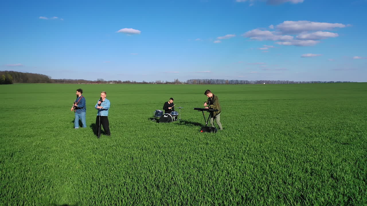 Boys' band plays music in the green field. Drone footage of the musicians first approaching them then distancing and flying aside. Aerial view.