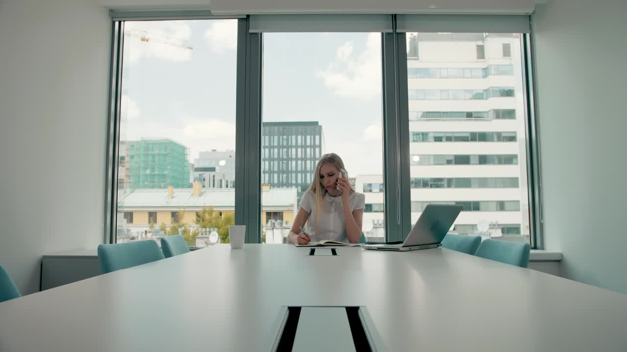 mujer seria trabajando en una habitación de oficina ligera. elegante mujer de negocios moderna con computadora portátil y papeles en una mesa larga en la sala de conferencias con luz haciendo una llamada telefónica