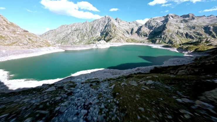 Gare di droni FPV su una riva lacustre sassosa e sorvolo a bassa quota sopra un lago glaciale color giada incorniciato da ripide creste elevate