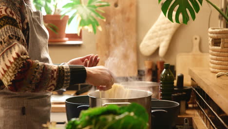 un homme en pull et en tablier cuisine des pâtes dans une casserole sur le poêle.