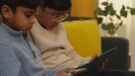 two young boys sitting on sofa at home playing games or streaming onto digital tablets 12