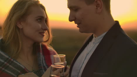 Young-married-couple-on-the-roof-hugging-and-drinking-red-wine-from-glasses-standing-dressed-in-plaid-and-admiring-the-beautiful-sunset-over-the-city..