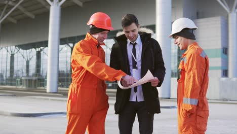 Inversionista-Del-Proyecto-Con-Traje-Negro-Examinando-El-Objeto-De-Construcción-Con-Trabajadores-De-La-Construcción-Con-Uniforme-Naranja-Y-Cascos.-Están-Revisando-Los-Dibujos-Juntos.