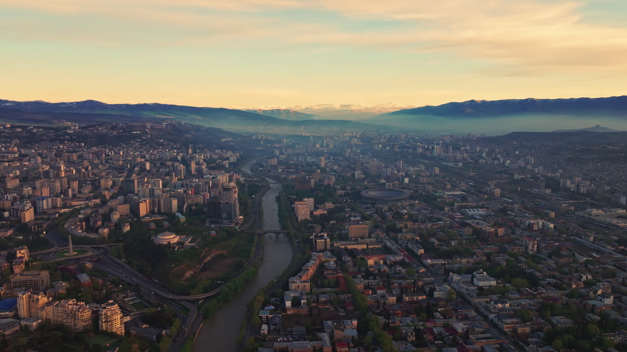 Aerial view of Tbilisi, Georgia, with the river winding through the city and mountains rising in the background, revealing urban texture, historic districts, and dramatic natural surroundings.