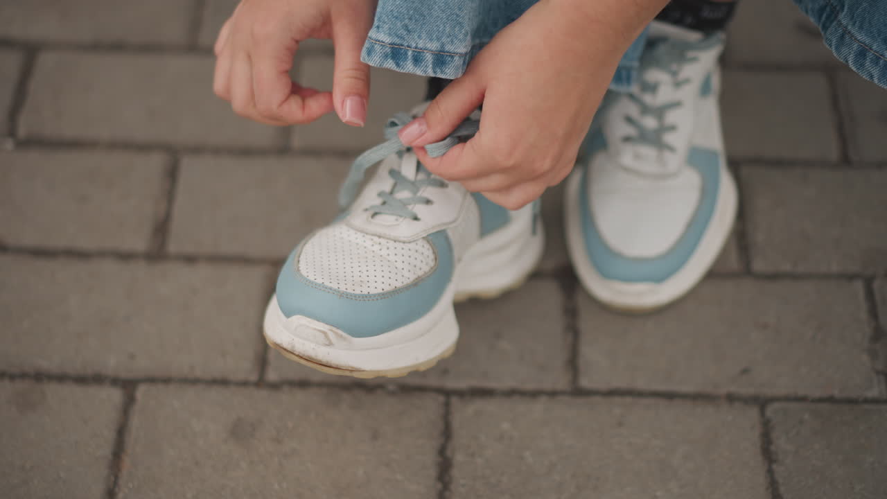 Una persona allaccia con cura scarpe da ginnastica blu. Primo piano di mani che fissano calzature sportive sul marciapiede cittadino. Un'immagine ravvicinata cattura le mani che legano scarpe sportive dai colori vivaci mentre il marciapiede cittadino attende il movimento.