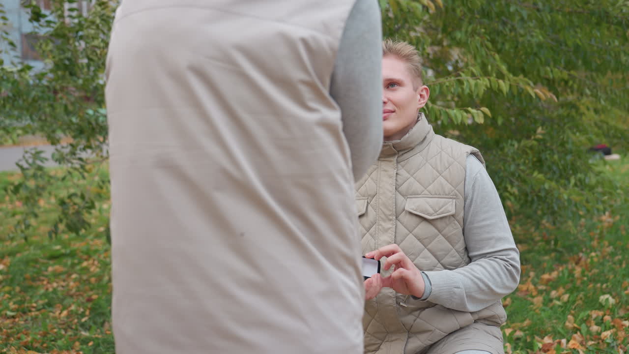 Heartfelt moment as man proposes to surprised woman who smiles with emotion while both wear matching outfits surrounded by green trees and fallen leaves