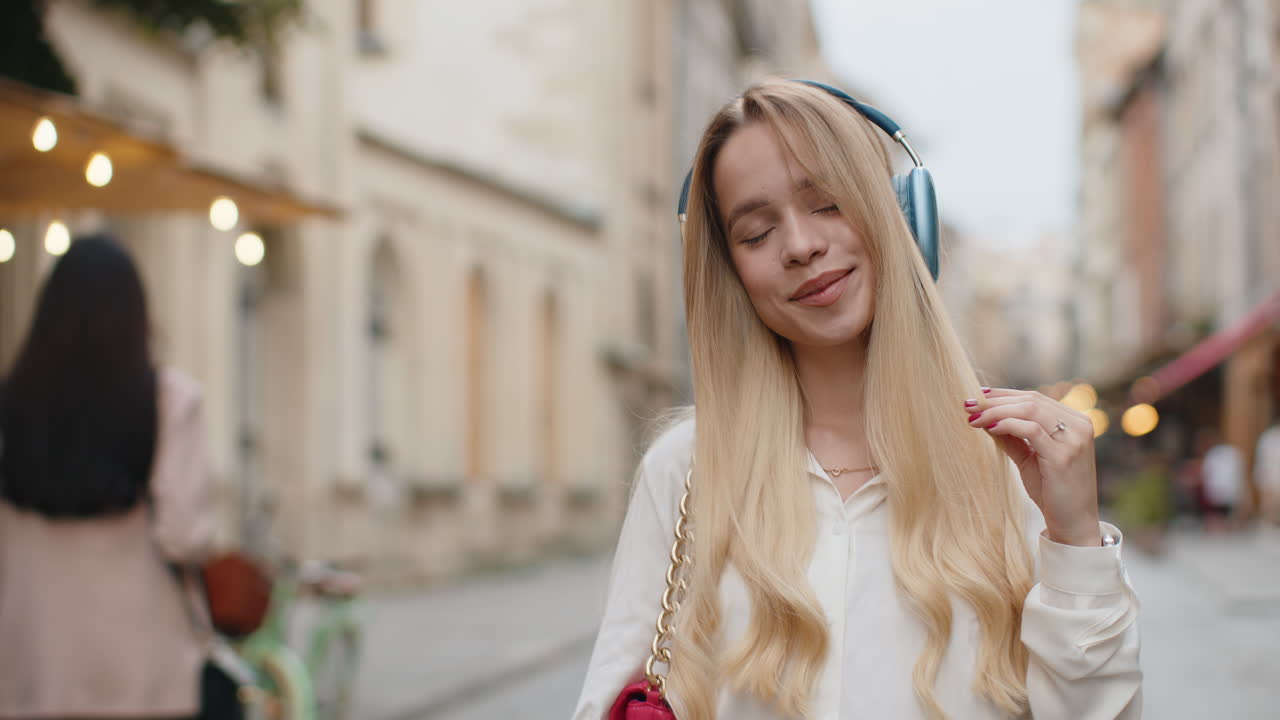 Happy young woman in wireless headphones choosing listening music dancing outdoors city street