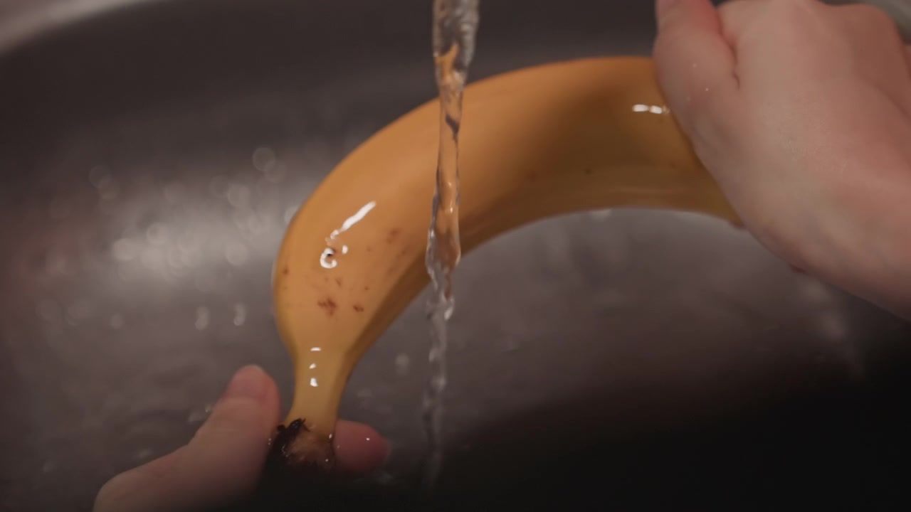Primer plano de manos lavando un plátano amarillo bajo el agua corriente en el fregadero. Las manos giran y frotan la fruta para limpiar todos los lados. El lavabo metálico del fregadero se muestra como detalle de fondo.