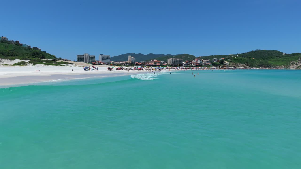 Bãi biển cát rộng với biển xanh ngọc tĩnh lặng tại Praia Grande, máy quay drone bay thấp theo sau những con hải âu. Arraial do Cabo, Rio de Janeiro, Brazil