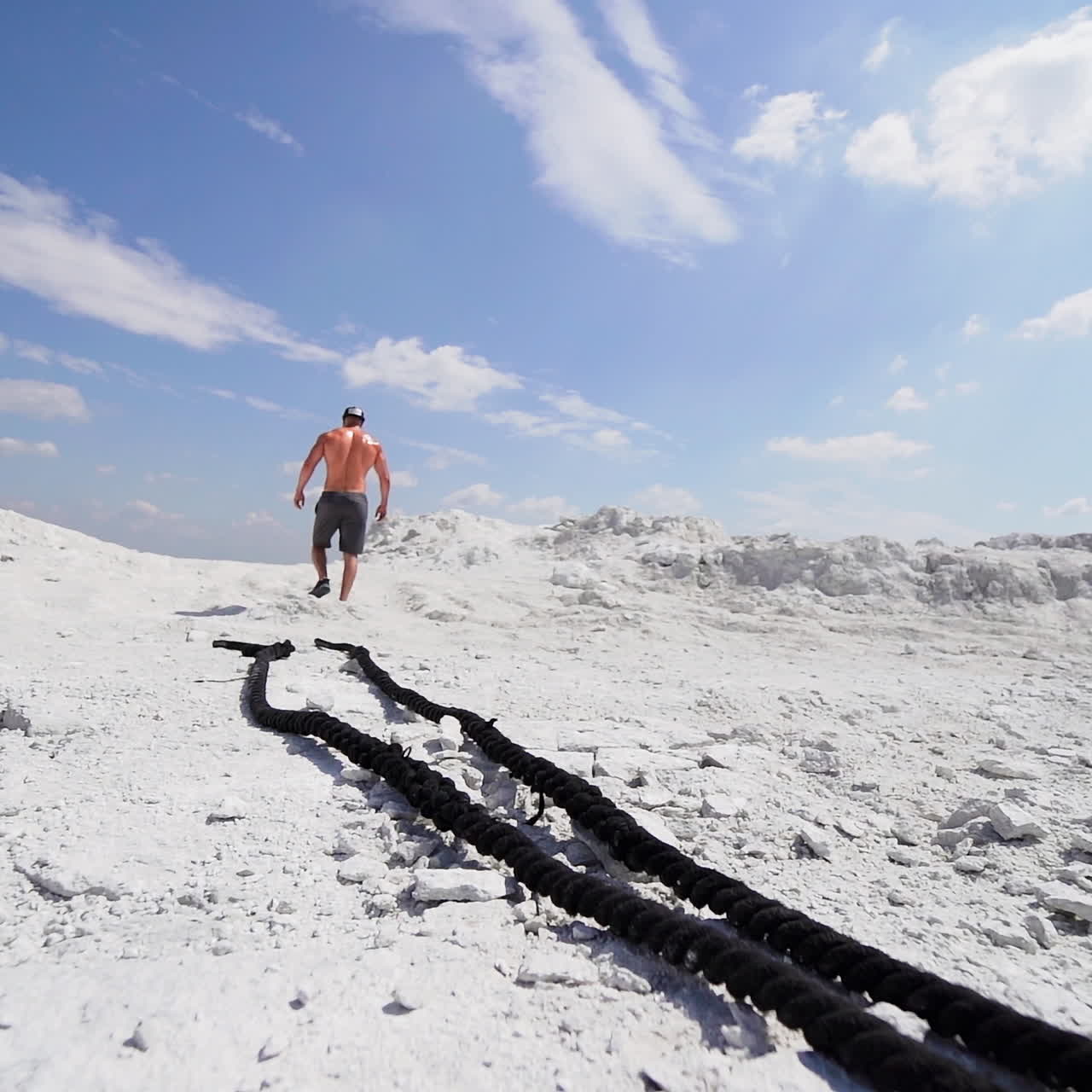 Shirtless man in shorts going up after his workout with ropes outdoors. Back view of a strong athlete rising on white mountain.