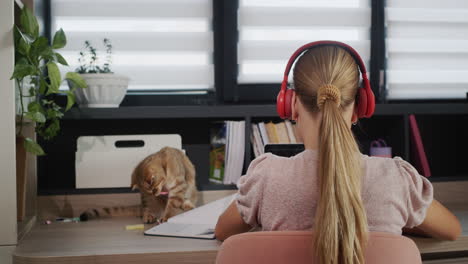 a teenage girl is studying at a laptop at home. she has headphones on, her pet helper sits next to her.