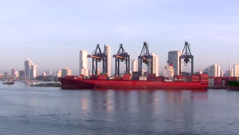 Grúas-Portuarias-Que-Mueven-Carga,-Con-Un-Barco-Rojo-En-Contraste-Con-Los-Edificios-Blancos