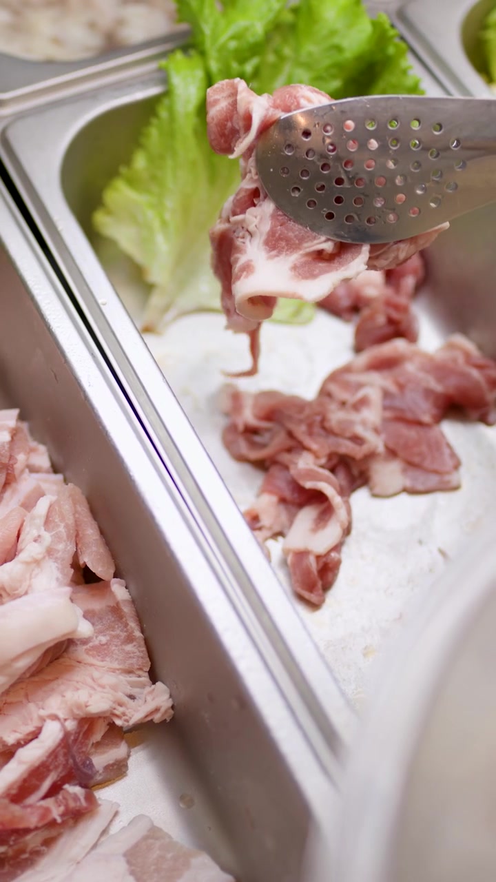 Tongs arrange raw sliced meat in stainless tray at buffet line, bright lighting, close-up view