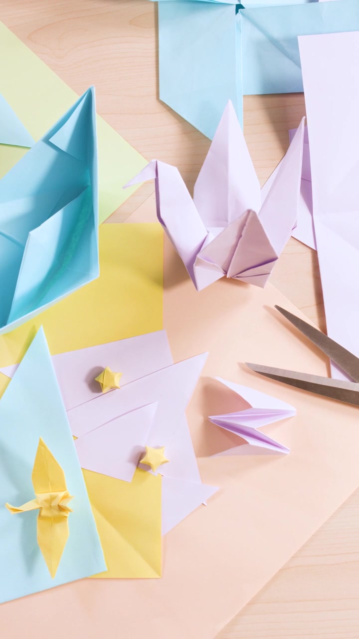 Overhead camera pans across pastel origami cranes, boats, and stars with crafting tools on wood