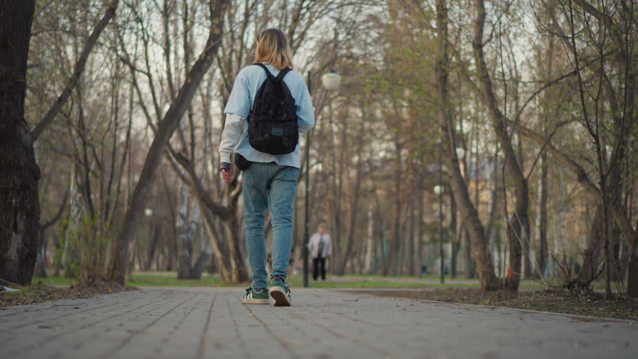 paseo tranquilo por el parque; una persona se aleja plácidamente por un sendero arbolado; un hombre con ropa informal se aleja por el camino del parque; un hombre caucásico con mochila da un paseo reflexivo por la tarde