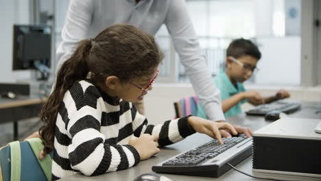 focused girl in eyeglasses sitting and doing task on computer while unrecognizable teacher looking at her