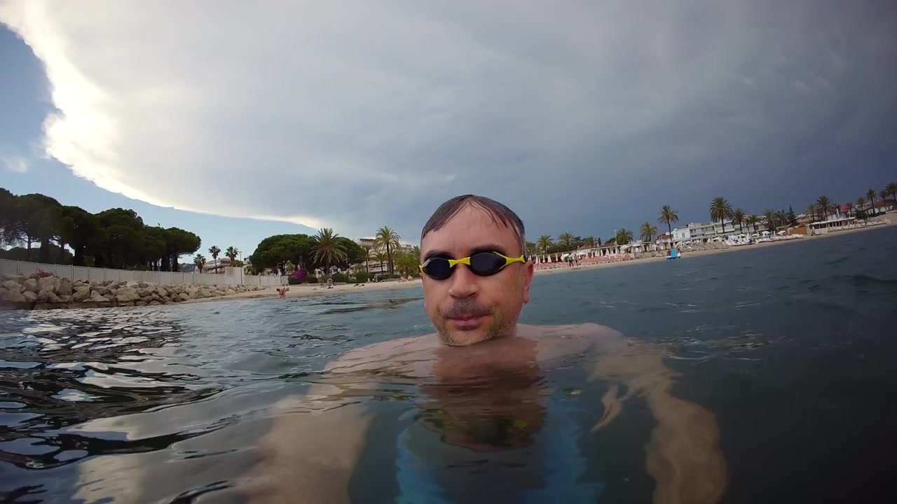 Riprese frontali di un nuotatore con occhialini che si muove in acque oceaniche calme vicino a una spiaggia tropicale sotto cieli nuvolosi e suggestivi.