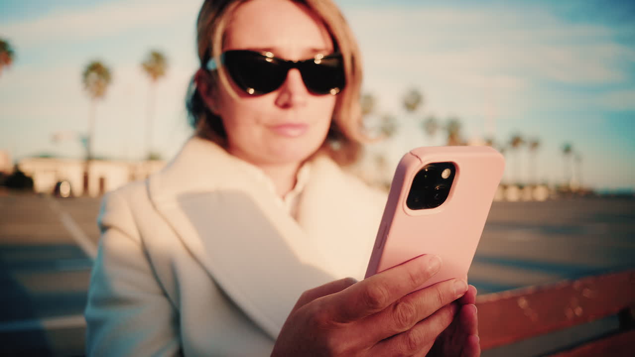 Stylish woman in a white coat looks at her smartphone while sitting on a wooden bench in a sunny parking area lined with palm trees