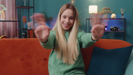 young woman smiling friendly at camera, waving hands gesturing hello, hi, greeting at home on sofa