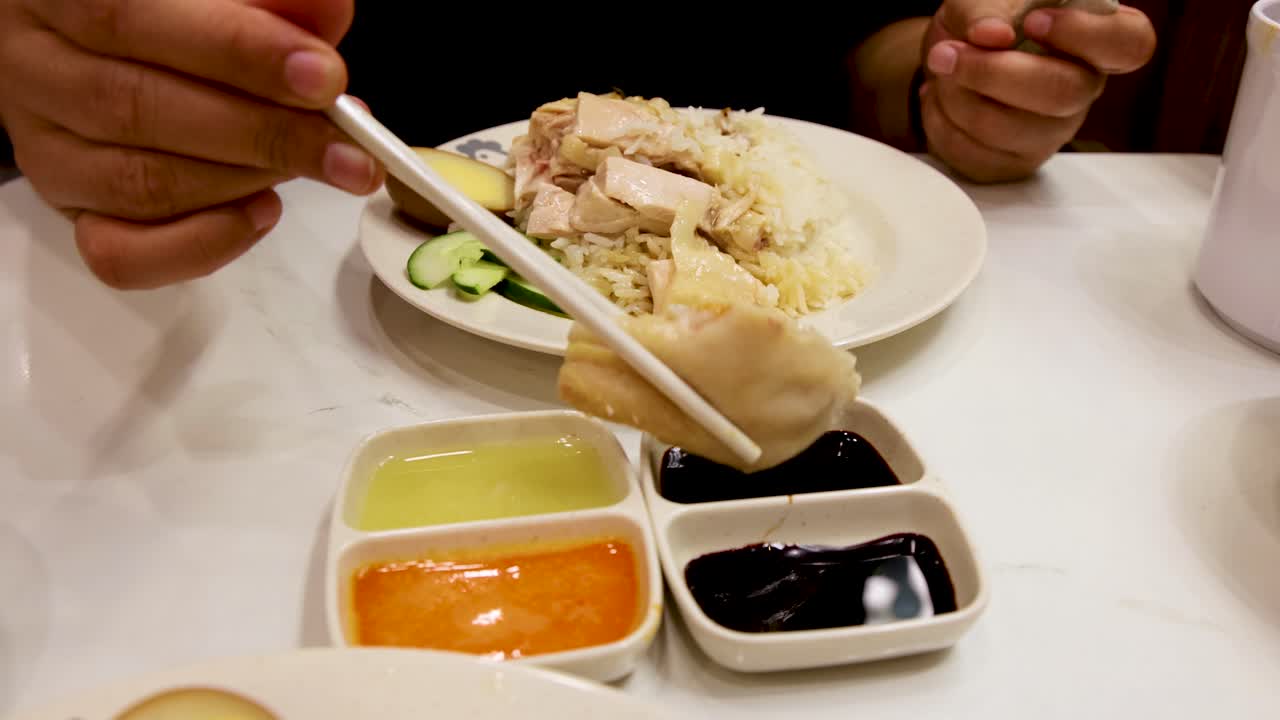 Hand uses chopsticks to dip a piece of steamed chicken into dark soy sauce beside rice, cucumber, and three dipping sauces on a restaurant table under bright lighting
