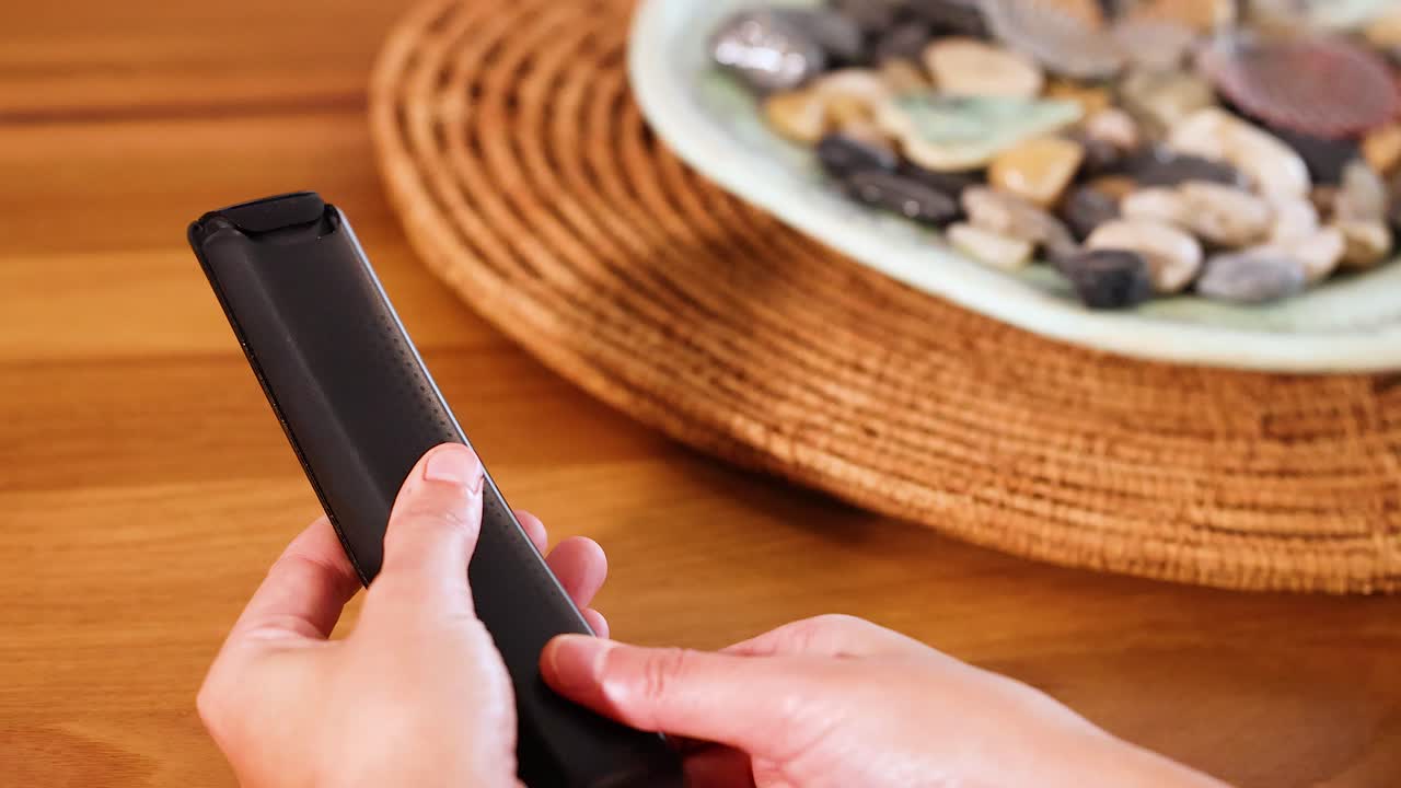 Hands replace batteries in a remote control on a wooden table with natural lighting and a decorative background