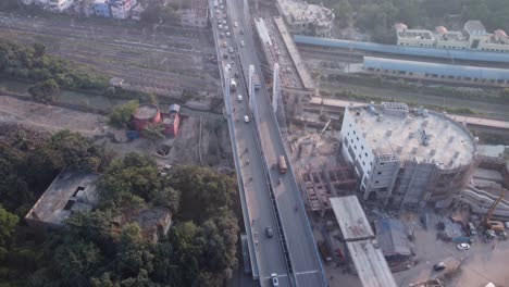 Toma-Aérea-Que-Revela-Lentamente-Los-Vehículos-Que-Cruzan-Majerhat-Sobre-La-Carretera-Del-Puente-Junto-Al-Nuevo-Sitio-De-Construcción-De-La-Línea-De-Metro-Y-La-Estación-De-Tren-Rodeada-Por-El-Paisaje-Urbano