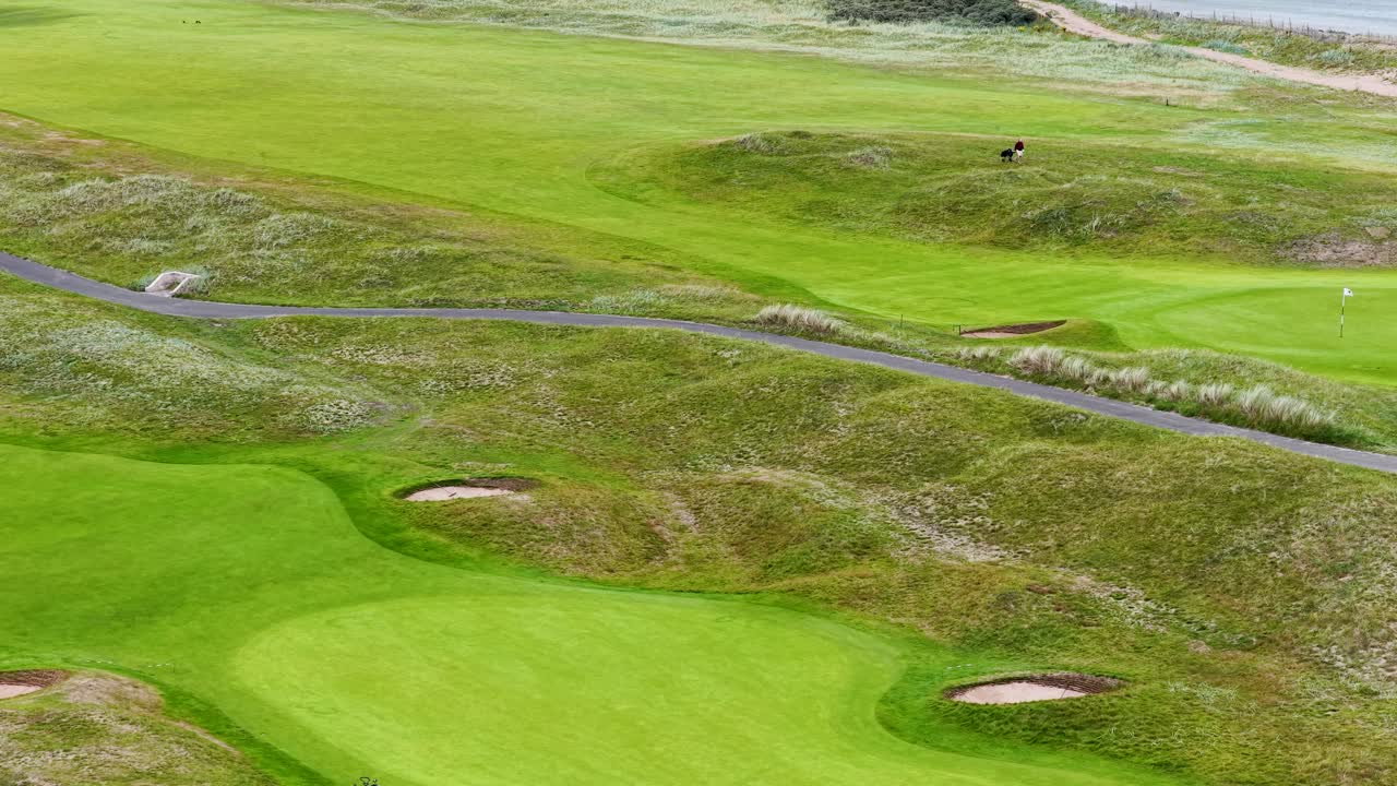 Drone glides above golfers playing on lush, coastal links course under soft daylight