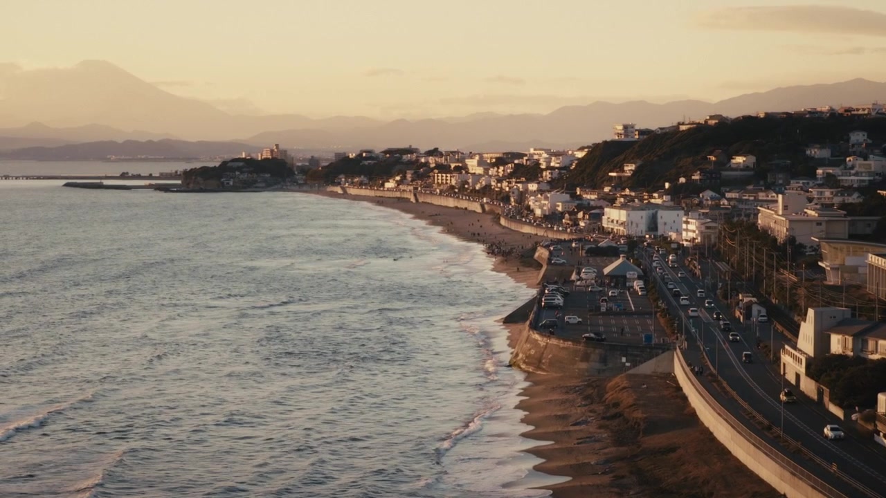 Tramonto aereo sulla tortuosa strada costiera e sulla spiaggia sabbiosa di Shichirigahama vicino a Kamakura, con edifici collinari, traffico di passaggio e la costa che si estende verso l'isola di Enoshima.