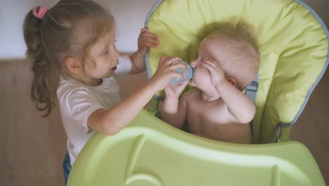 sister strokes baby giving water to brother in highchair