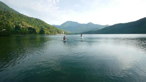 two people in distance using stand up paddle board on lake amidst the mountains during early morning sunrise, filmed as wide establishing shot