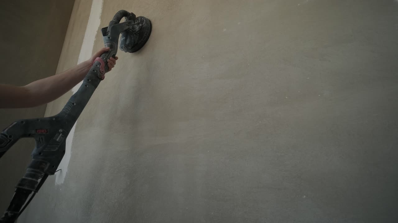 Close-up image of a hand holding a power sander against a freshly plastered wall, focusing on the tool and surface texture