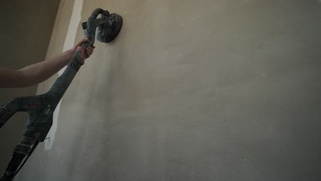 close-up image of a hand holding a power sander against a freshly plastered wall, focusing on the tool and surface texture