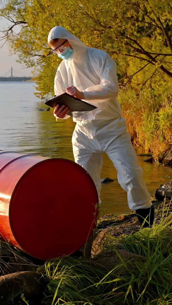 Scientist in hazmat gear rolls oil drum by lakeside during field inspection for pollution, vertical