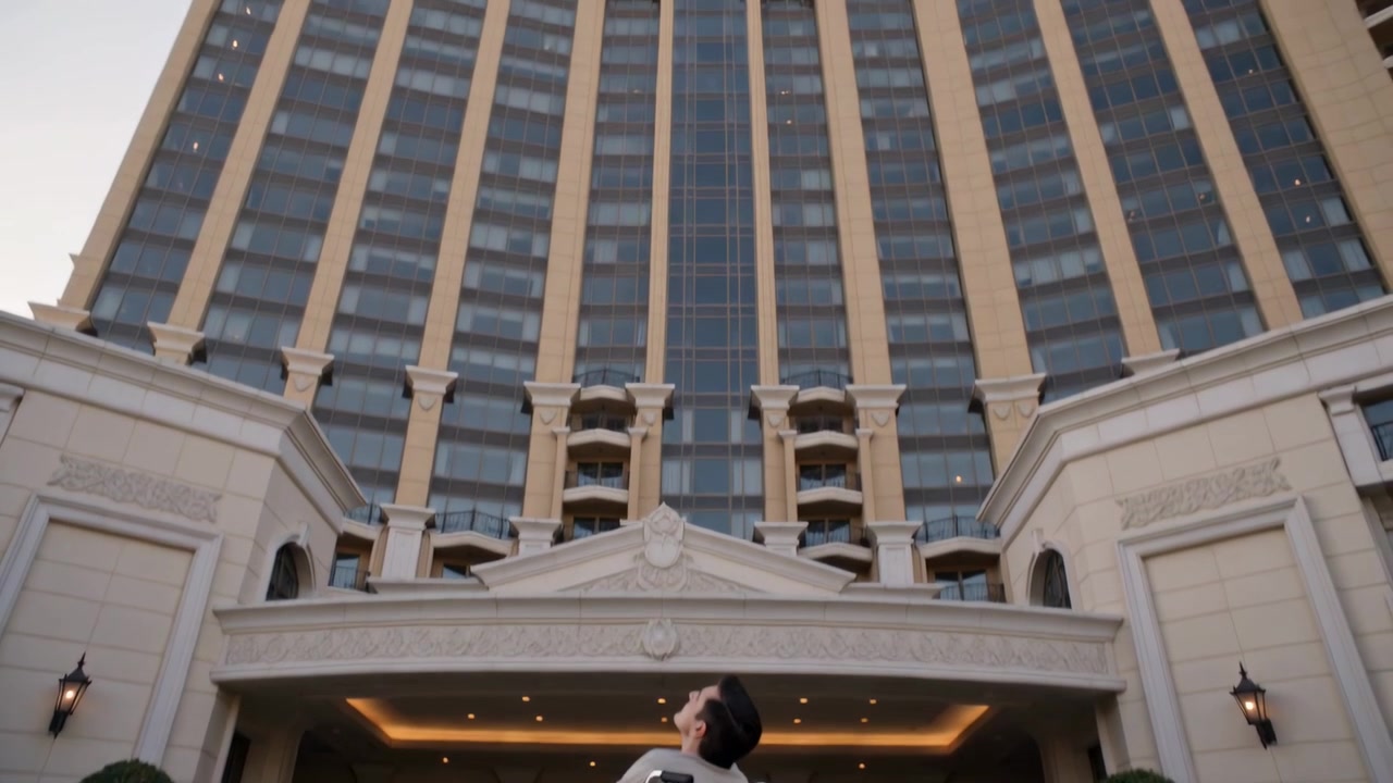 La telecamera si inclina verso l'alto, un uomo in sedia a rotelle con camicia chiara e jeans ammira la facciata dell'hotel all'arrivo