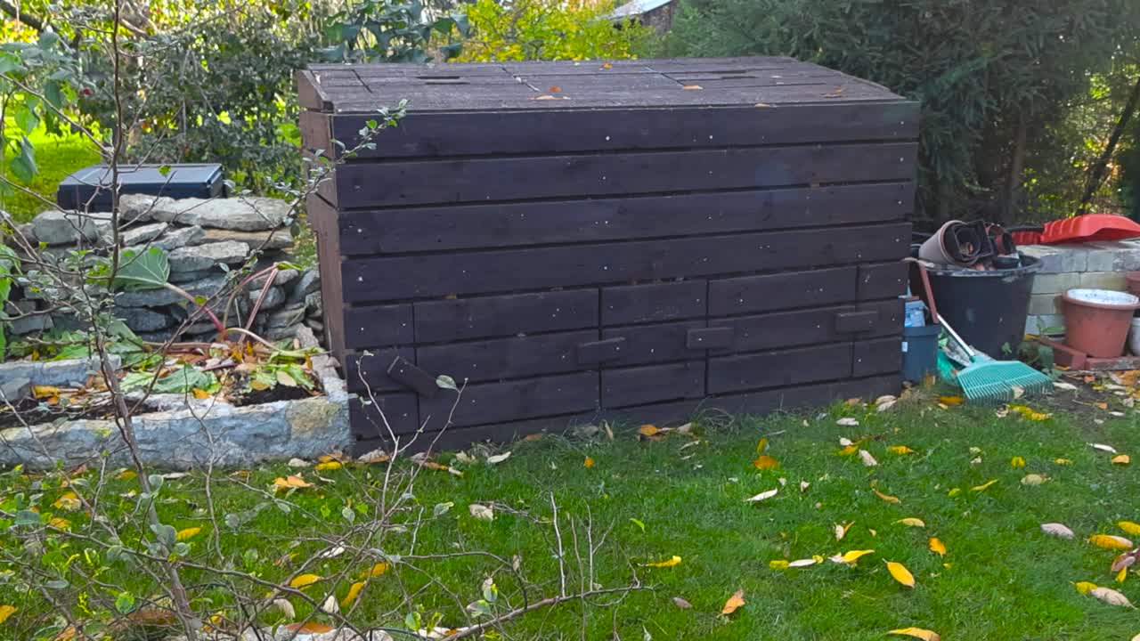 Brown DIY wooden composter or compost bin in a autumn garden with green mowed grass lawn and yellow fallen leaves during a cloudy day with limestone garden beds on the side. Sun is shining in the back