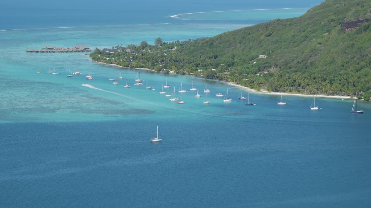 View of sailboats, overwater bungalows, beaches, palm trees, coral reefs and the ocean on a sunny day in a small green tropical pacific island from Magic Mountain, Moorea, French Polynesia.