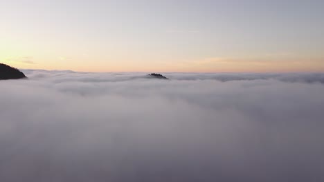 drone flies over a layer of clouds where the top of a mountain protrudes during a sunrise