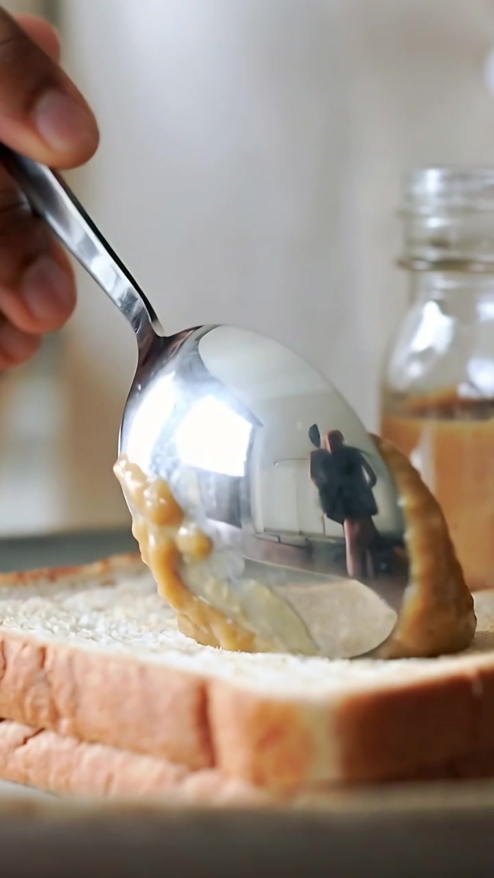 Vertikal video: Hånd dypper ske, skovler lysebrunt smørepålæg fra krukke på brød ved køkkenbordet, iført skjorte