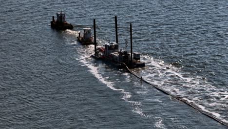 an aerial view of two tugboats, and a small barge dredging a bay on long island, new york on a sunny day