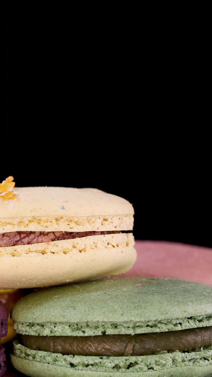 A close-up view of a hand delicately placing assorted macarons atop a stack, with vibrant colors and dramatic studio lighting against a black backdrop