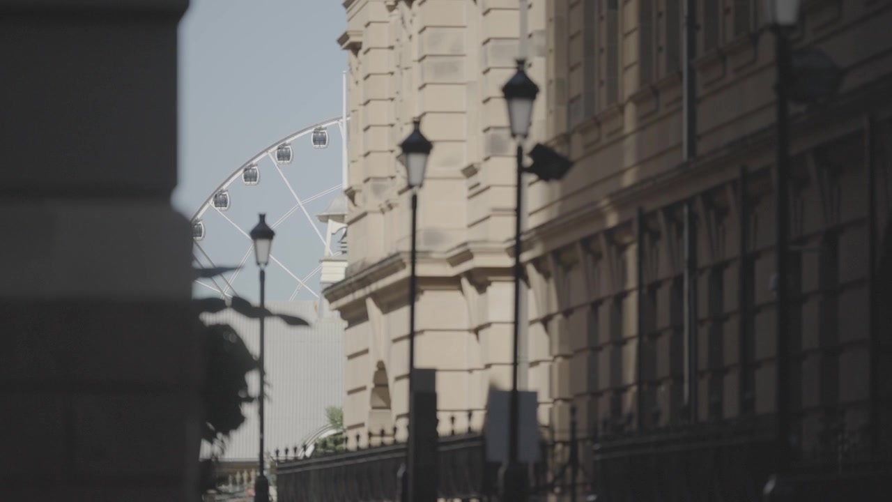 Una vista a nivel de calle muestra un edificio histórico de arenisca bordeado de farolas, con una noria visible a lo lejos.