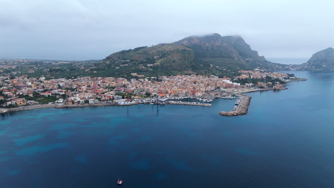 Drone shot of Porticello’s vibrant waterfront and hills with a small red rescue vessel returning to port from the site of the sunken Bayesian yacht nearby