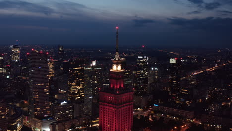 Forwards-fly-around-top-of-old-high-rise-building-with-cityscape-in-background.-Red-lit-Palace-of-Culture-and-Science.-Warsaw,-Poland
