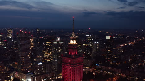 Vorwärts-Fliegen-Um-Die-Spitze-Des-Alten-Hochhauses-Mit-Stadtbild-Im-Hintergrund.-Rot-Beleuchteter-Kultur--Und-Wissenschaftspalast.-Warschau,-Polen