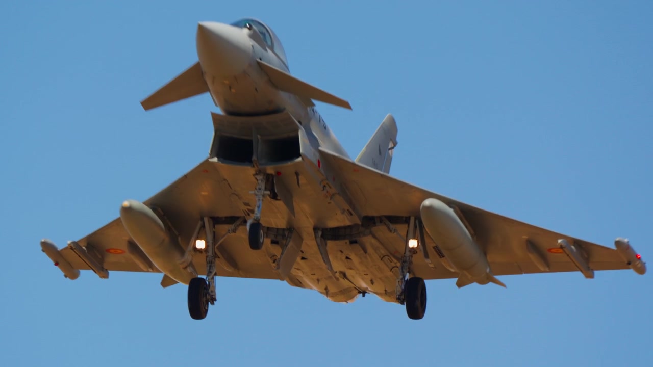 Military fighter jet approaching for landing with its landing gear extended, flying against a clear blue sky in slow motion