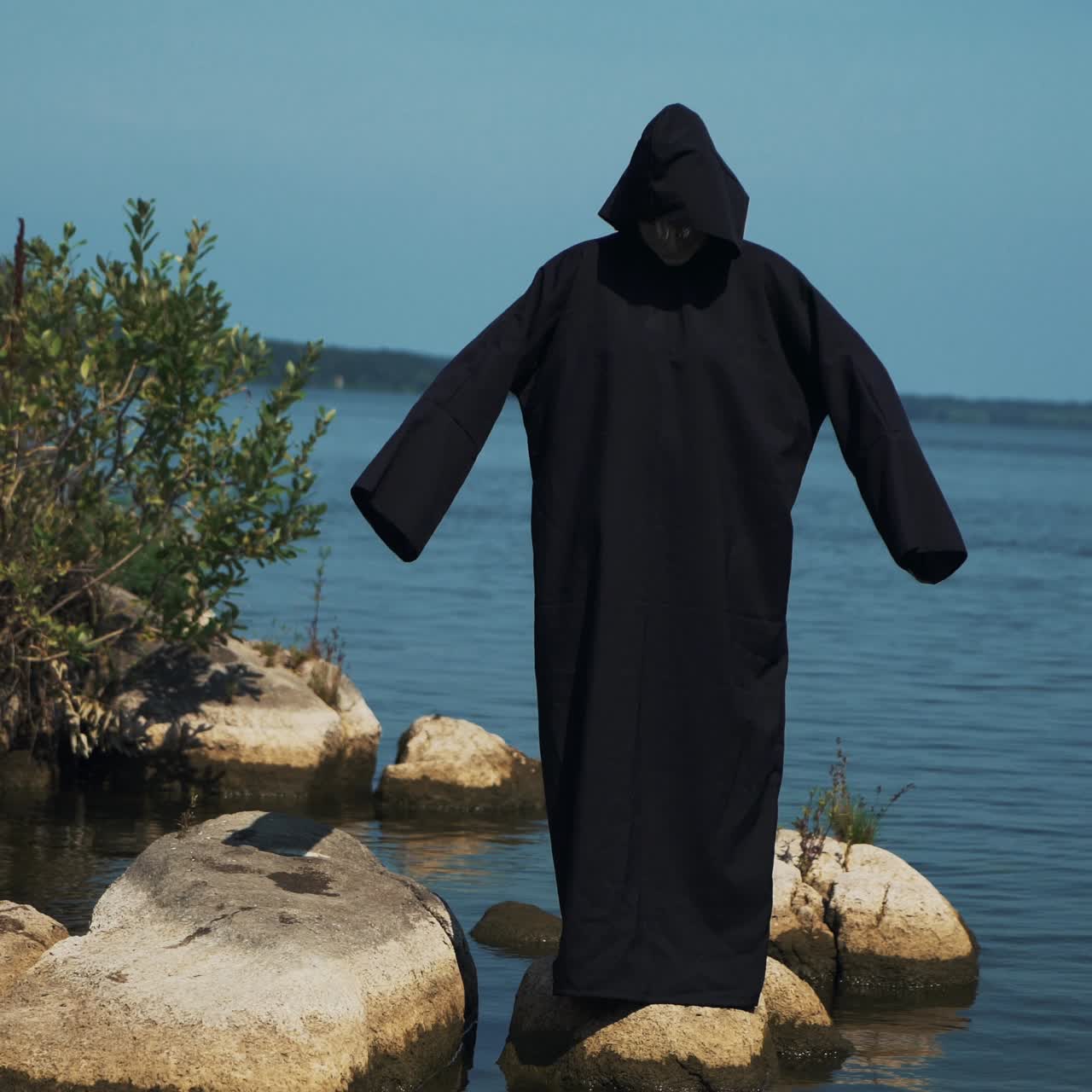 Woman devil in black cape and hood on stones near the river. Halloween costume.