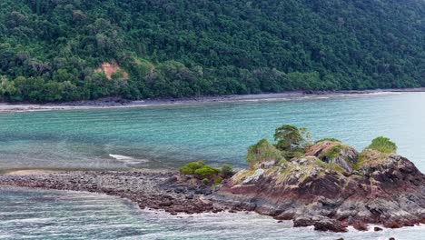 drone camera glides over a small rocky island with lush greenery, turquoise water, and forested coastline in soft daylight, revealing tranquil coastal scenery